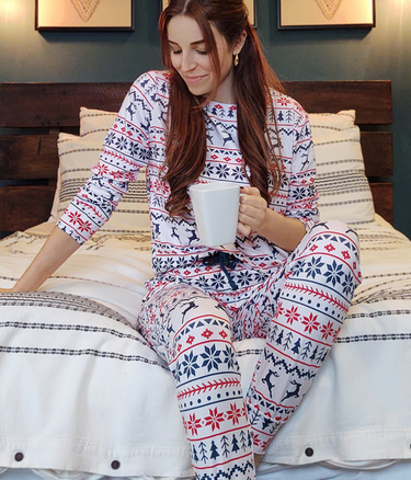 Woman wears the White Christmas Pyjamas whilst sitting on her bed with a hot drink.